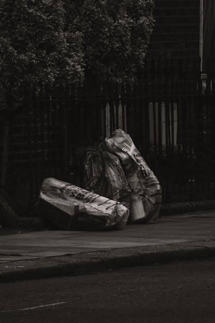 Two black plastic bags of waste, visibly crumpled and tied at the top, rest on a paved sidewalk near a black metal fence and a large leafy bush in an outdoor urban setting. One bag appears to contain 