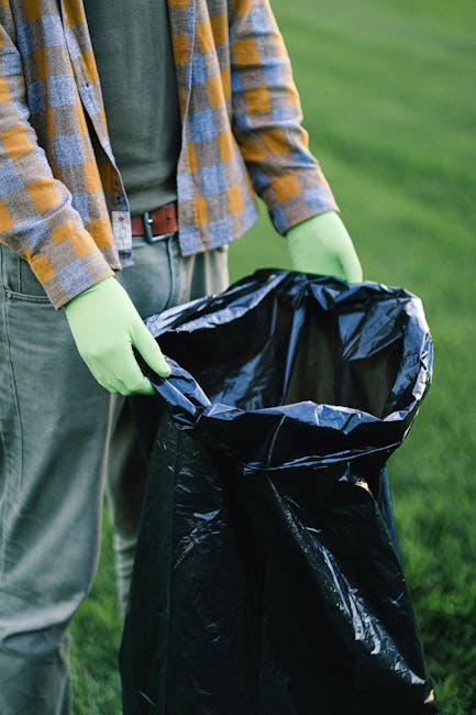 Two black plastic bags of waste, visibly crumpled and tied at the top, rest on a paved sidewalk near a black metal fence and a large leafy bush in an outdoor urban setting. One bag appears to contain 