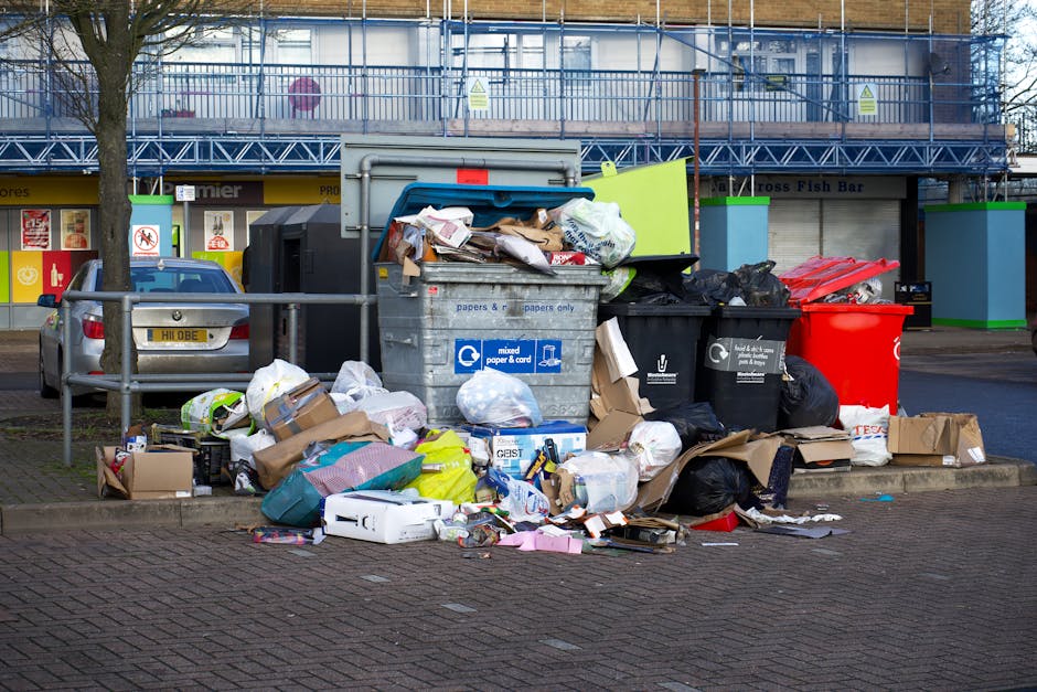 A black plastic wheelie bin with the label 'St. Johns' standing on a paved sidewalk beside a residential street at night. The bin is filled with various types of rubbish, including cardboard boxes and